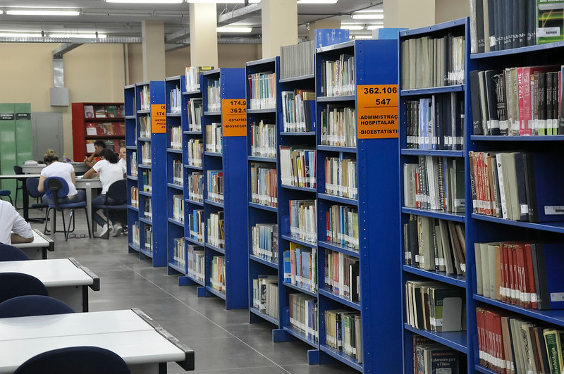 Imagem: foto mostra uma biblioteca da UFC onde aparecem várias estantes de livros e, ao fundo, uma mesa com algumas estudantes sentadas