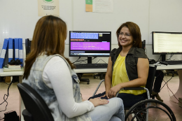 Imagem: foto de uma servidora cadeirante conversando com outra sentada numa cadeira de costas, atrás da cadeirante, há uma bancada com um computador ligado exibindo o programa DOXVOX