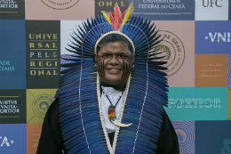 Foto do estudante Benício Pitaguary, licenciado em Geografia pela UFC, trajando beca de formatura e um cocar comprido com penas azuis