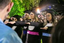 Imagem: foto dos concluintes vestidos de beca com braço direito estendido para o juramento dos formandos