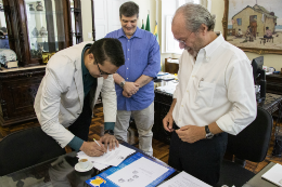 Imagem: foto em que aparecem o novo diretor da biblioteca assinando o termo de posse, o reitor ao lado direito e o prof. Maurício ao fundo