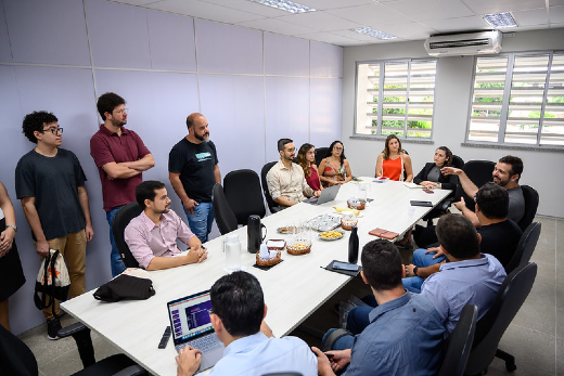 Imagem: grupo de pessoas reunidas em mesa de reunião branca, em sala com janela, algumas em pé.