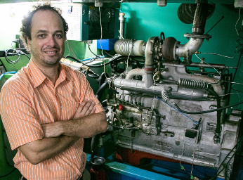 Imagem: Foto mostra o professor André Bueno, da cintura para cima, com braços cruzados, em frente a um motor automotivo (Foto: Ribamar Neto/UFC Informa)