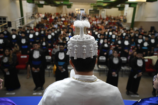 Imagem: Imagem mostra o reitor de costas, trajando vestes talares. Ao fundo, olhando para a câmara, jovens formandos e suas famílias aguardam a solenidade (Foto: Viktor Braga/UFC Informa)