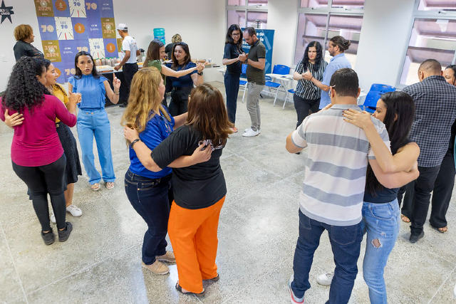Imagem: A imagem registra uma atividade realizada no campus de Quixadá durante a programação do Mês do Servidor 2024. O espaço amplo e iluminado recebe um grupo de servidores — docentes e técnico-administrativos — que participam de uma oficina de forró. Organizados em pares, eles praticam passos básicos, em postura de condução e acompanhamento, com expressões de concentração e descontração. Ao fundo, é possível ver cadeiras azuis alinhadas e cartazes que reforçam o caráter institucional e celebrativo do momento.