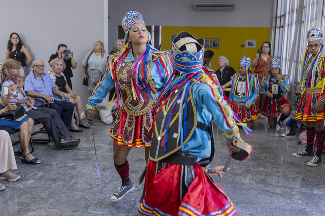Imagem: Em um salão iluminado, dois dançarinos em trajes coloridos e tradicionais realizam uma coreografia animada diante de um público atento. Outros integrantes, também fantasiados, acompanham ao fundo. (Foto: Guilherme Silva/UFC Informa)
