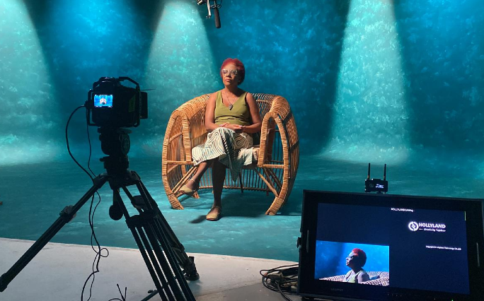 Após a exibição do filme haverá roda de conversa (Demitri Túlio/Divulgação) Imagem: Mulher negra sentada em uma cadeira de palha fala, dentro de um estúdio, em frente a um fundo que simula as cores do mar. Em frente a ela, há uma câmera e um computador (Foto: Demitri Túlio/Divulgação)