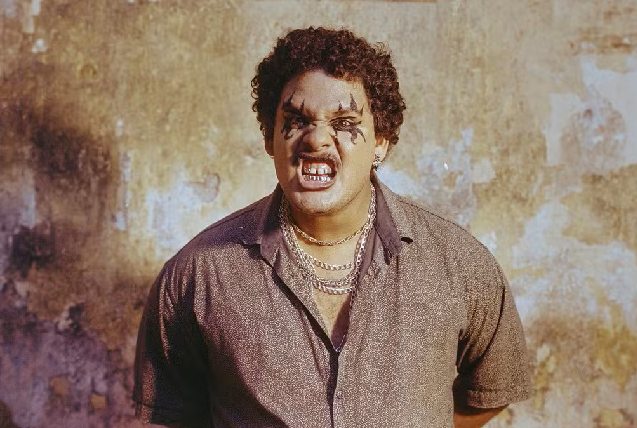 Imagem: A imagem mostra um homem com expressão agressiva, gritando com os dentes à mostra e o rosto marcado por uma maquiagem escura ao redor dos olhos. Ele veste uma camisa simples e várias correntes douradas, posando diante de uma parede desgastada pelo tempo (Foto: Jorge Silvestre/Divulgação)