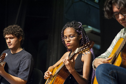 Imagem: foto de três jovens, dois rapazes e uma moça ao centro tocando violão