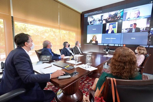 Participantes da reunião em volta da mesa olhando para o telão de videochamada com outros participantes