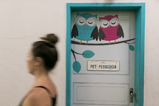 Imagem: Mulher de pele branca e cabelos pretos em frente à porta do PET Pedagogia