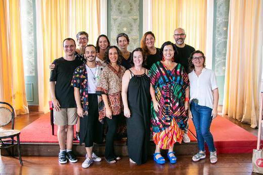 Imagem: professores em pé posam para foto em ambiente de cortinas e janela ao fundo