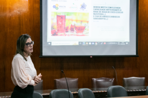 Imagem: Foto mostra professora em pé, com camisa branca e saia preta, a frente de uma parede onde está sendo projetada uma apresentação sobre bebida mista feita a partir de água de coco (foto: Ribamar Neto/UFC Informa)