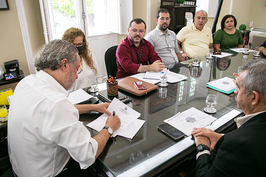 Imagem: foto do reitor Cândido Albuquerque sentado à cabeceira de uma mesa retangular no Gabinete assinando um documento. Ao redor da mesa, estão sentados outros participantes da reunião 