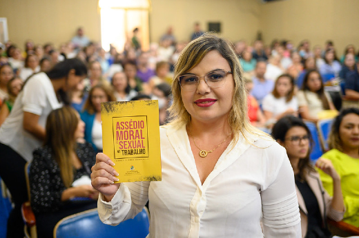 Imagem: mulher loira de óculos com blusa branca segura cartilha Assedio Moral em capa amarela e ao fundo uma plateia de auditório repleta de pessoas. 