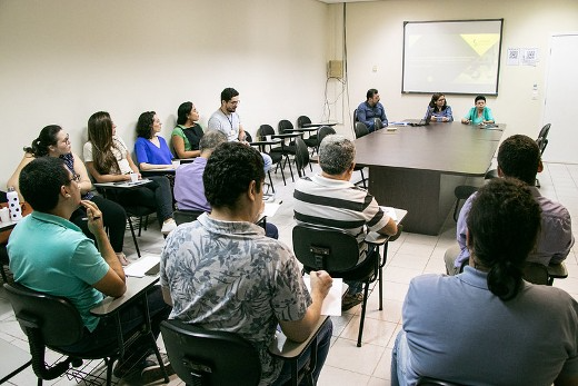 Imagem: foto de uma sala de aula com pessoas sentadas nas cadeiras. Ao fundo, três pessoas estão sentadas de frente para as pessoas