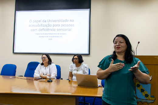 Imagem: mesa de auditório e painel ao fundo, duas mulheres de blusa branca sentadas na mesa e uma mulher de cabelos escuros e blusa verde em pé fazendo interpretação de libras