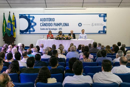 Plateia de costas, com pessoas sentadas nas poltronas azuis do auditório Cândido Pamplona, identificado ao fundo com uma placa grande, à frente da qual colocou-se uma mesa longa com toalha branca, à qual estão sentados quatro servidores