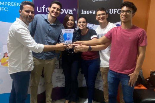 Equipe de seis pessoas (dois homens à esquerda, duas mulheres ao centro e dois homens à direita) posa para foto com as mãos estendidas ao centro, segurando um troféu em formato de placa, nas cores azul e branco 