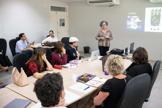 Imagem: Diana Azevedo aparece de pé, junto à cabeceira de uma longa mesa branca, ocupada por várias pessoas. Atrás dela aparece uma projeção de uma apresentação