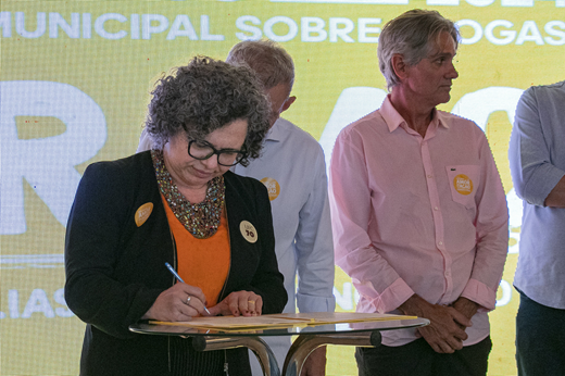 Imagem: A vice-reitora Diana Azevedo assina o termo de cooperação com a Prefeitura de Fortaleza; ela traja blazer preto e está posicionada em frente a um telão. (Foto: Álvaro Graça Jr./ UFC Informa)