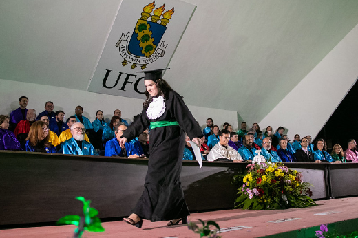 Imagem: mulher caminha com beca na frente de mesa formada