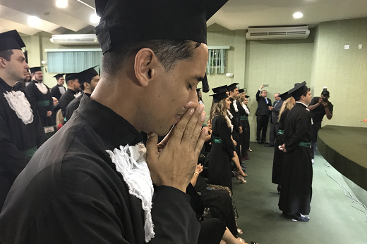 Imagem: foto de jovens de beca em pé em um auditório em posição de juramento