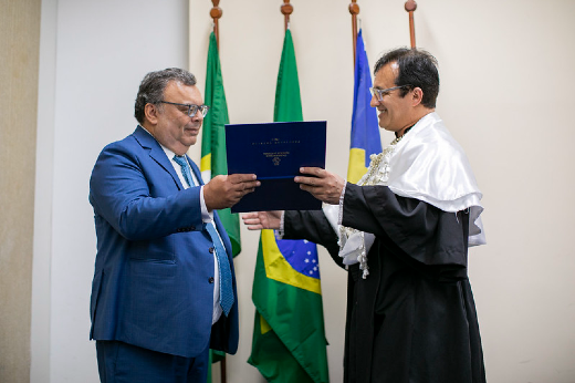 Imagem: Homem de terno recebe título das mãos de homem vestido com roupas de reitor, preta e branca, com bandeiras ao fundo