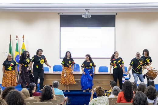 Imagem: A imagem mostra um grupo de oito pessoas em pé, dançando e sorrindo em um palco. Eles vestem camisetas pretas com desenhos coloridos de formas geométricas e elementos do sol. Algumas das pessoas estão usando saias longas e coloridas, enquanto um dos homens à direita toca um tambor. Ao fundo, há uma tela de projeção, bandeiras do Brasil e outra amarela com um brasão, além de cadeiras azuis. O público está sentado em cadeiras de cor clara, assistindo à apresentação.