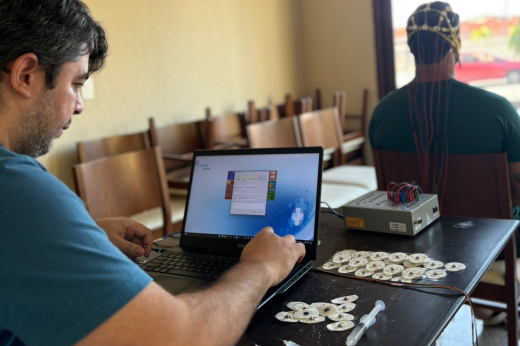 Imagem: Em primeiro plano, psicólogo em frente a um computador, acompanhando gráfico de um eletro. Ao fundo, atleta com eletrodos conectados à cabeça (Foto: Letícia Clemente/Divulgação)