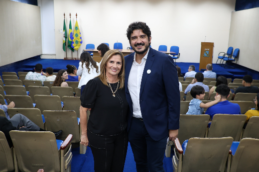 Imagem: A imagem mostra duas pessoas, um homem e uma mulher, sorrindo para a câmera. Eles estão no centro de um auditório com várias fileiras de cadeiras marrons vazias ao redor, exceto por algumas pessoas ao fundo. No palco à frente, há bandeiras, incluindo a do Brasil, e um púlpito de madeira. O homem está usando um terno azul escuro e camisa branca, enquanto a mulher usa uma blusa preta e calça combinando. O ambiente tem um tom formal, possivelmente relacionado a um evento institucional ou cerimonial.