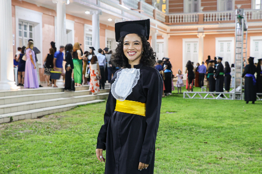 Imagem: mulher de cabelo preto e cacheado posa sorrindo para foto, vestida com uma beca de formatura preta, acompanhada por uma faixa amarela e um chapéu de graduação. Ao fundo, aparece parte da fachada do andar térreo do prédio da reitoria da UFC e vários outros formandos também vestindo beca. A imagem foi tirada em um evento de formatura ao ar livre, sobre um gramado. 