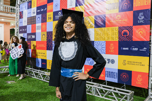 Imagem: pessoa vestida com uma beca de formatura preta, acompanhada por uma faixa azul e um chapéu de graduação. A pessoa está sorrindo e posando para a câmera, com uma mão na cintura. Ao fundo, há um painel colorido com o logo da Universidade Federal do Ceará (UFC) e outros símbolos relacionados à formatura, junto com o texto "Formei!" e o brasão da universidade. A imagem parece ter sido tirada em um evento de formatura ao ar livre, sobre um gramado. No canto esquerdo, outra pessoa segura um cartaz com o texto "EU <3 UFC