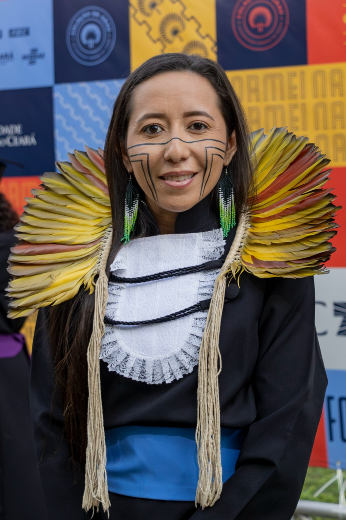 Imagem: jovem mulher de cabelos pretos e lisos, cocar no pescoço e pintura com traços pretos no rosto posa para foto vestindo beca e faixa azul