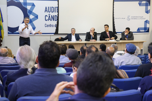 Imagem: homem de cabelos grisalhos encaracolados e curtos, de camisa social branca e calça jeans aparece falando ao microfone sobre um palco em um auditório, ao lado de uma mesa comprida atrás da qual aparecem sentados quatro homens, todos de camisa social e blazer. Na frente deles, olhando para eles, aparecem várias pessoas de costas para a câmera, sentados nas poltronas do auditório