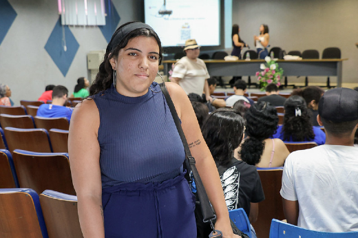 Imagem: A imagem mostra uma jovem em primeiro plano, usando uma blusa sem mangas azul-escura e uma bolsa de ombro preta. Ela está em pé em um auditório, com fileiras de cadeiras e algumas pessoas ao fundo. No palco, duas mulheres parecem estar terminando uma apresentação. A jovem sorri levemente, e no fundo é possível ver detalhes da decoração e arranjos de flores no palco.