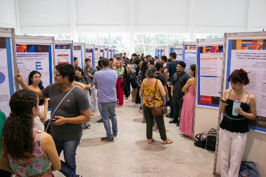 Ambiente de congresso acadêmico com diversas pessoas de pé em um corredor entre painéis de pôsteres científicos. Algumas pessoas conversam em grupos sobre os trabalhos expostos, enquanto outras observam os pôsteres ou fazem anotações. O local é bem iluminado com luz natural, e os pôsteres abordam diferentes temas acadêmicos, com textos e gráficos visíveis. A cena transmite um clima de troca de conhecimento e aprendizado