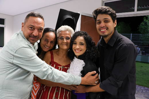 Imagem: família reunida em uma celebração, todos sorrindo e abraçados, com uma mulher ao centro usando uma beca, destacando um momento especial de conquista