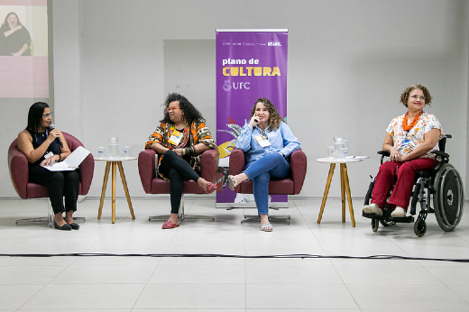 Imagem: A imagem mostra quatro mulheres sentadas em cadeiras dispostas em linha, participando de um evento em um ambiente interno iluminado. Ao fundo, há um banner roxo com o texto "plano de CULTURA UFC", relacionado à Universidade Federal do Ceará (UFC), acompanhado de elementos gráficos.  À esquerda, uma mulher de cabelos pretos e lisos usa óculos e veste roupa preta. Ela segura folhas de papel e parece estar moderando ou conduzindo a conversa. A segunda mulher, com cabelos cacheados volumosos, veste uma blusa estampada em tons de laranja, preto e branco, e está sorrindo enquanto cruza as pernas. A terceira mulher, com cabelos ondulados e claros, usa uma camisa azul clara e jeans. Ela parece estar falando ou respondendo, com expressão descontraída. À direita, uma mulher em cadeira de rodas, de cabelos curtos e cacheados, veste uma blusa branca estampada e calça vermelha. Ela sorri enquanto participa da discussão. Entre as cadeiras, há pequenas mesas com copos e jarras de vidro. O ambiente sugere um painel ou debate focado em temas culturais ou diversidade.