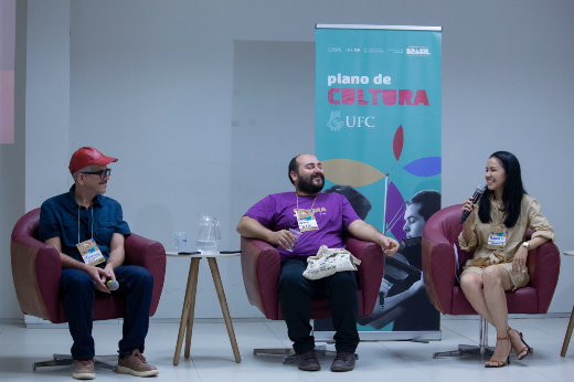 Imagem: A imagem mostra três pessoas sentadas em cadeiras de formato moderno, em um ambiente interno iluminado. Atrás deles, há um banner com o texto "plano de CULTURA UFC", acompanhado do logotipo da Universidade Federal do Ceará (UFC) e de elementos gráficos coloridos.  À esquerda, está um homem usando um boné vermelho, camisa escura e óculos. Ele tem um crachá pendurado no pescoço e está olhando para a pessoa no centro. No centro, há um homem de barba e cabelo curto, vestindo uma camiseta roxa com o texto "CULTURA". Ele segura um tênis branco no colo enquanto sorri. À direita, uma mulher de cabelos lisos e pretos veste um vestido bege e segura um microfone enquanto sorri, aparentemente falando ou interagindo. Entre as cadeiras, há uma pequena mesa com um copo e uma jarra de vidro. O ambiente sugere um evento ou painel relacionado a cultura e educação.