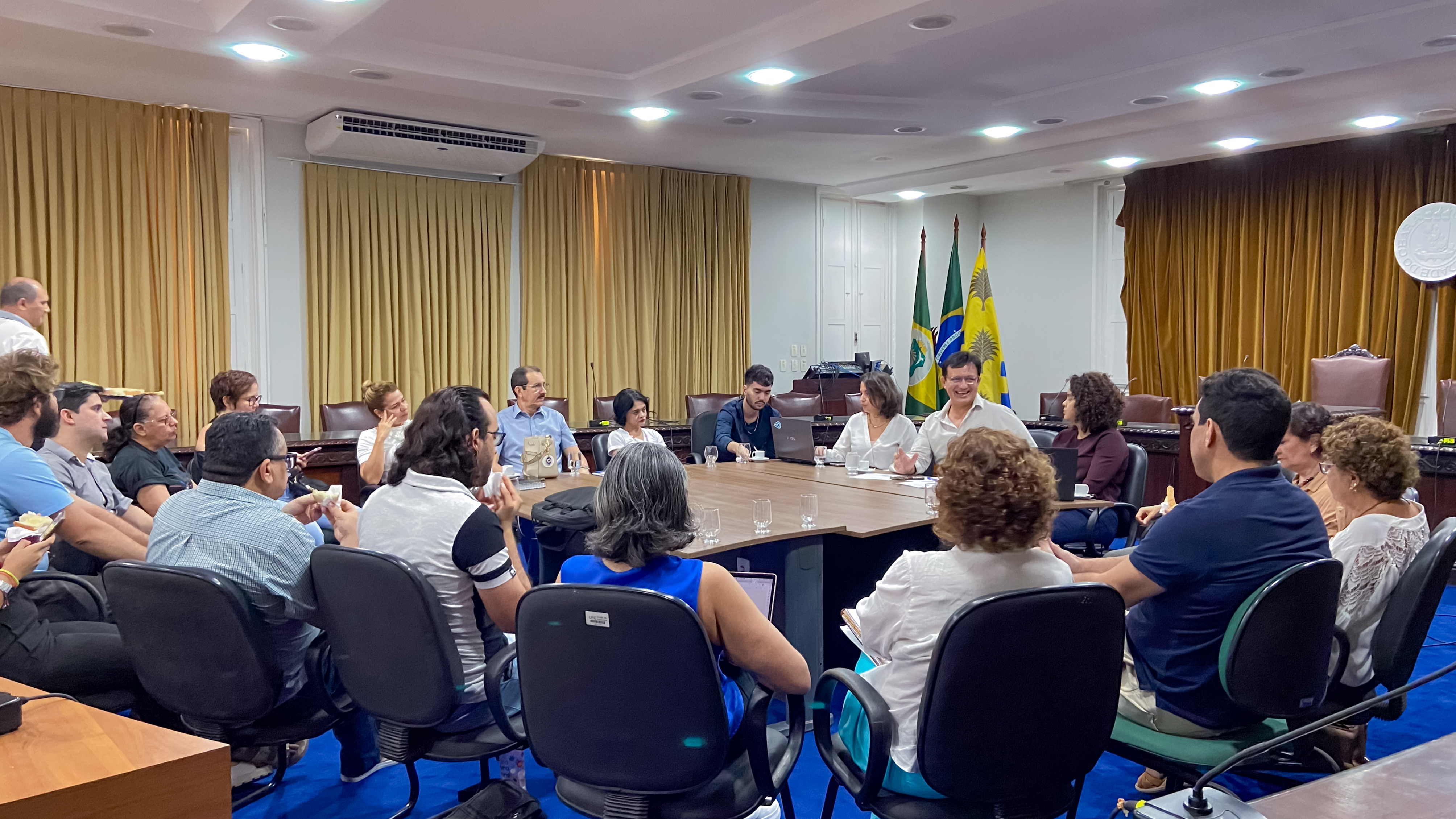 Imagem: Por volta de 20 pessoas de diferentes gêneros e idades estão reunidas e sentadas em volta de uma mesa, dentre essas pessoas, está o reitor Custódio Almeida.