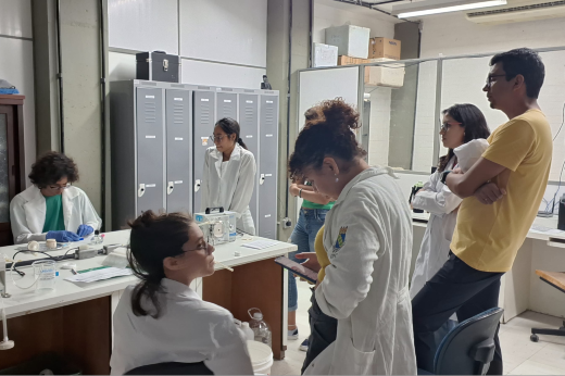 Imagem: grupo de pessoas em um laboratório. Algumas estão usando jalecos brancos e outras roupas casuais. Elas estão em atividades diversas: uma pessoa trabalha em um microscópio, outras estão de pé observando ou conversando. O ambiente contém armários, bancadas de trabalho e equipamentos de laboratório