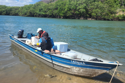 Imagem: uma pequena embarcação azul e prata chamada 'TAINHA', ancorada em um rio próximo à margem. Duas pessoas estão a bordo, vestindo coletes salva-vidas e chapéus, manuseando equipamentos e caixas de isopor. Ao fundo, há vegetação densa e água límpida sob um céu azul.