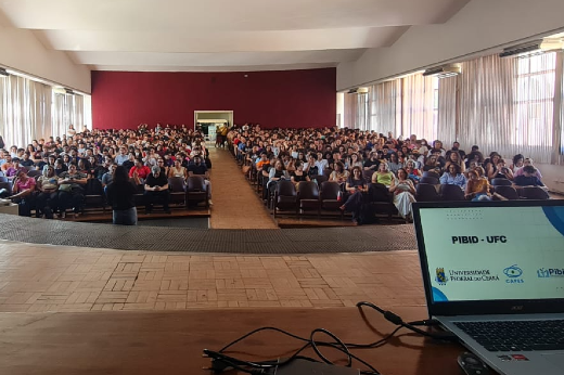 Imagem: A imagem mostra um auditório amplo, com muitas pessoas sentadas em fileiras de cadeiras, assistindo a uma apresentação. O auditório possui paredes brancas com janelas altas nas laterais, que deixam a luz natural entrar. No fundo, há uma parede de cor vermelha, e à frente, um palco de madeira com um notebook posicionado sobre uma mesa. Na tela do notebook, é possível ler "PIBID - UFC" e os logos da Universidade Federal do Ceará (UFC) e do programa PIBID. A perspectiva é do palco, com visão para a plateia, destacando a grande quantidade de pessoas no local.