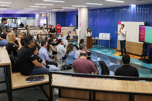 Imagem: um jovem apresentador, vestindo camisa branca e calça jeans, está de pé na frente de uma plateia, segurando um microfone e um controle remoto. Ele gesticula enquanto apresenta slides exibidos em uma tela grande ao seu lado. A plateia, composta por homens e mulheres de diferentes idades e estilos, está sentada em arquibancadas e cadeiras, prestando atenção. O espaço tem um design moderno, com paredes azuis, iluminação clara e um ambiente colaborativo. Ao fundo, há uma mulher de pé perto de algumas impressoras e um grupo de pessoas observando a apresentação