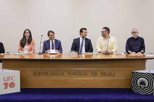 Imagem: Cinco pessoas estão sentadas atrás de uma mesa de madeira em um auditório da Universidade Federal do Ceará (UFC). A mesa tem o nome da instituição gravado na frente. À esquerda, uma mulher de óculos veste um blazer salmão e uma blusa branca. É a vice-diretora da Editora UFC, Juliana Diniz. Ao lado dela, dois homens usam terno e gravata, um deles, sorrindo e segurando papéis, é o prefeito de Fortaleza, Evandro Leitão. Ao lado dele, aparece o reitor da UFC. Custódio Almeida. O quarto participante veste uma camisa amarela de mangas compridas e conversa com um dos homens de terno. É o diretor da Editora UFC. Cavalcante Júnior. O último homem, à direita, tem barba branca, usa óculos e veste uma camisa preta. É o escritor Lira Neto.Na frente da mesa, há dois painéis com o logotipo da UFC e a inscrição "70 anos". O fundo da sala é claro e neutro.