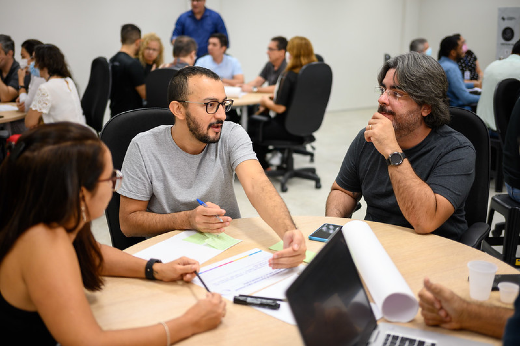 Imagem: A imagem mostra um grupo de pessoas em um ambiente interno participando de uma reunião ou atividade colaborativa. Em primeiro plano, três pessoas estão sentadas em volta de uma mesa redonda. Um homem com barba e óculos, vestindo uma camiseta cinza, gesticula enquanto fala com os outros. À sua direita, um homem com cabelos grisalhos e óculos, vestindo uma camisa preta, escuta atentamente, com a mão no queixo. À esquerda, uma mulher com cabelo castanho escuro preso e óculos participa da conversa. Na mesa, há papéis, canetas e um notebook aberto. Ao fundo, outras pessoas estão sentadas em grupos, também interagindo. O ambiente parece ser um escritório ou sala de reuniões com cadeiras giratórias e iluminação clara.