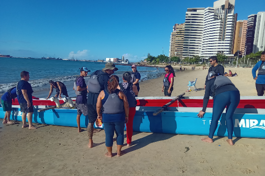 Imagem: grupo de pessoas reunidas em uma praia ensolarada, ao lado de uma grande canoa azul com detalhes vermelhos. A canoa tem a palavra "MIRA" visível na lateral. Os participantes vestem roupas esportivas, muitos com coletes salva-vidas e mochilas. Alguns estão segurando remos, enquanto outros parecem estar organizando a embarcação. No fundo, há um mar calmo com alguns barcos visíveis ao longe, prédios altos à beira-mar e algumas pessoas caminhando na areia. O céu está azul e sem nuvens, indicando um dia de clima bom.