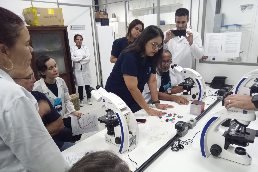 Imagem: Um grupo de pessoas em um laboratório, reunidas em torno de uma mesa com microscópios. Uma mulher de óculos e camisa azul escura está no centro, inclinada sobre a mesa, com as mãos sobre pequenos objetos coloridos espalhados. Outras pessoas, algumas de jaleco branco e outras de camisa azul, observam atentamente e participam da atividade. Ao fundo, um homem de jaleco branco registra o momento com um celular, e uma mulher também de jaleco branco está encostada em um móvel de madeira com os braços cruzados. O ambiente tem prateleiras com materiais de laboratório e equipamentos científicos.