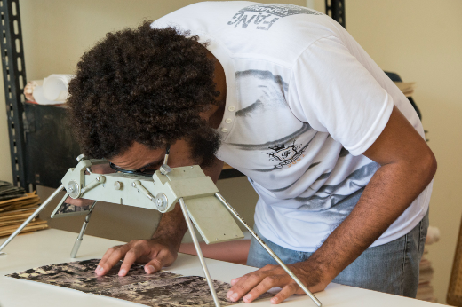 Imagem: Um homem com cabelo cacheado e barba, vestindo uma camiseta branca com estampa cinza e calça jeans, está curvado sobre uma mesa analisando uma imagem com a ajuda de um estereoscópio. O estereoscópio é um equipamento metálico com pernas ajustáveis e lentes binoculares, utilizado para observação de imagens em três dimensões, comumente em análises cartográficas e geográficas. O homem posiciona as mãos sobre a imagem enquanto observa atentamente através do aparelho. Ao fundo, há prateleiras com documentos e fichários organizados, sugerindo um ambiente de pesquisa ou arquivo técnico.